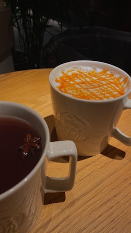 Two white ceramic mugs sit on a wooden table. One contains a cappuccino with a layer of frothy milk topped with a criss-cross pattern of orange-colored powder. The other mug holds a dark tea with a star anise floating on top. The background is slightly blurred with dark hues, possibly indicating a cafe setting.