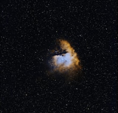A celestial scene with a vibrant nebula surrounded by a field of stars. The nebula exhibits a blend of blue and orange hues, suggesting a rich interaction of gases. The surrounding space is dotted with numerous small stars against a dark backdrop.