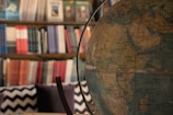 A large globe is prominently displayed in the foreground, showcasing detailed geographical features. In the background, a bookshelf filled with an array of colorful books, organized in a gradient of colors, adds depth to the setting. The room has a cozy and scholarly vibe.