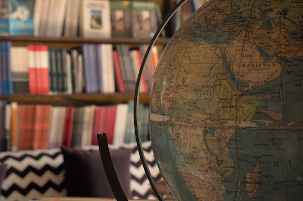 A large globe is prominently displayed in the foreground, showcasing detailed geographical features. In the background, a bookshelf filled with an array of colorful books, organized in a gradient of colors, adds depth to the setting. The room has a cozy and scholarly vibe.
