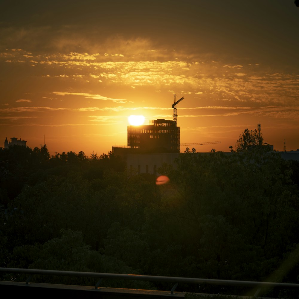 the sun is setting behind a building with a crane in the foreground