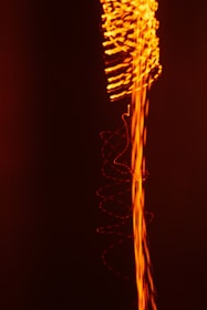 A dynamic composition featuring swirling and wavy lines of bright orange light against a deep black background, creating an impression of energy and movement.