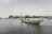A traditional wooden fishing boat with a long tail engine travels through calm waters. There are a few people aboard, and in the background, lush green trees line the shore. The sky is overcast with pale clouds, creating a tranquil atmosphere.