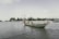 A traditional wooden fishing boat with a long tail engine travels through calm waters. There are a few people aboard, and in the background, lush green trees line the shore. The sky is overcast with pale clouds, creating a tranquil atmosphere.