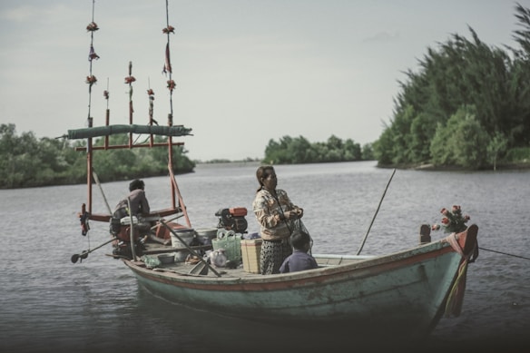 A small wooden boat is gently floating on a calm river, with two adults and a child on board. The boat is equipped with fishing gear and decorated with floral elements. Lush greenery lines the riverbanks under a partly cloudy sky.