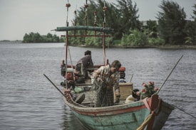 A small wooden fishing boat is floating on a calm river or lake, surrounded by lush greenery. Two people are on board; one is adjusting a fishing net while the other is seated towards the stern. The boat is decorated with floral elements, and fishing equipment is scattered around.