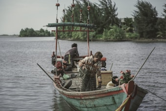 A small wooden fishing boat is floating on a calm river or lake, surrounded by lush greenery. Two people are on board; one is adjusting a fishing net while the other is seated towards the stern. The boat is decorated with floral elements, and fishing equipment is scattered around.