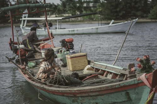 A small wooden fishing boat with two people working on it floats on a calm body of water. The boat has red and turquoise accents with various fishing equipment, nets, and containers onboard. In the background, another similar boat is seen further along the river, with lush green trees lining the shore.