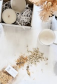 A serene arrangement featuring a candle box with shredded packing paper, an open candle with a wick, and another candle placed on a white surface. Dried flowers are scattered around, with a branded package labeled 'Calendula Petals & Rose.' Soft artificial flowers in a vase are partially visible.