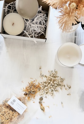 A serene arrangement featuring a candle box with shredded packing paper, an open candle with a wick, and another candle placed on a white surface. Dried flowers are scattered around, with a branded package labeled 'Calendula Petals & Rose.' Soft artificial flowers in a vase are partially visible.