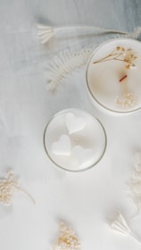 Two white candles are placed on a soft, textured surface. One of the candles has a smooth top with a wooden wick and small dried flowers. The other candle contains heart-shaped designs. Delicate white dried flowers and ferns are scattered around them, creating an elegant and serene composition.