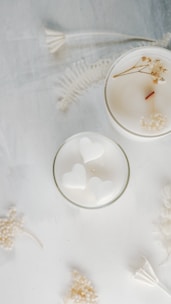Two white candles are placed on a soft, textured surface. One of the candles has a smooth top with a wooden wick and small dried flowers. The other candle contains heart-shaped designs. Delicate white dried flowers and ferns are scattered around them, creating an elegant and serene composition.