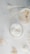 Two white candles are placed on a soft, textured surface. One of the candles has a smooth top with a wooden wick and small dried flowers. The other candle contains heart-shaped designs. Delicate white dried flowers and ferns are scattered around them, creating an elegant and serene composition.