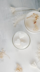 Two white candles are placed on a soft, textured surface. One of the candles has a smooth top with a wooden wick and small dried flowers. The other candle contains heart-shaped designs. Delicate white dried flowers and ferns are scattered around them, creating an elegant and serene composition.