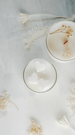 Two white candles are placed on a soft, textured surface. One of the candles has a smooth top with a wooden wick and small dried flowers. The other candle contains heart-shaped designs. Delicate white dried flowers and ferns are scattered around them, creating an elegant and serene composition.