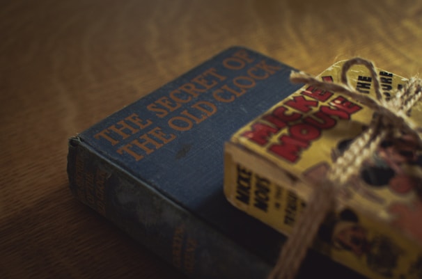 A vintage hardcover book titled 'The Secret of the Old Clock' lies on a wooden surface next to a bundle of comic books tied with rough string. The book cover is dark blue with bold orange lettering, while the comics feature vibrant, colorful illustrations.