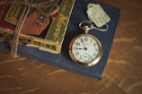An antique pocket watch with a gold casing rests on top of a closed, navy blue book. The watch's face displays bold black numbers and ornate black hands. Beside it, a vintage comic book is bound with rustic twine, featuring colorful illustrations on its cover. A small white tag with handwritten details is tucked under the watch chain.