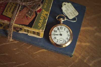 An antique pocket watch with a gold casing rests on top of a closed, navy blue book. The watch's face displays bold black numbers and ornate black hands. Beside it, a vintage comic book is bound with rustic twine, featuring colorful illustrations on its cover. A small white tag with handwritten details is tucked under the watch chain.