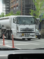 A large white tanker truck is parked on a city street, with two orange traffic cones placed nearby. The background features a modern building under construction, with scaffolding and fresh greenery. A few cars are visible around the truck, and the photo appears to be taken from inside another vehicle.