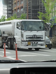 A large white tanker truck is parked on a city street, with two orange traffic cones placed nearby. The background features a modern building under construction, with scaffolding and fresh greenery. A few cars are visible around the truck, and the photo appears to be taken from inside another vehicle.
