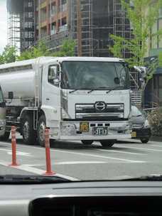 A large white tanker truck is parked on a city street, with two orange traffic cones placed nearby. The background features a modern building under construction, with scaffolding and fresh greenery. A few cars are visible around the truck, and the photo appears to be taken from inside another vehicle.