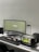 A modern, minimalist workspace featuring a curved monitor displaying the text 'ecorium Natural Decor'. A mechanical keyboard with a black, white, and red keys is placed on a dark desk mat. To the right of the workspace, there is a small, well-maintained terrarium with plants and moss. Other elements include a sleek black desk lamp, a mouse, a trackpad, and a small black speaker.