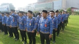 A group of people standing in lines on a grassy field wearing blue patterned uniforms and black hats. They appear to be organized in a formation and have ID badges on their chests. In the background, there is a large building with glass windows and a sign displaying 'RITA Super Mall'. Trees and other vegetation are visible along the perimeter.