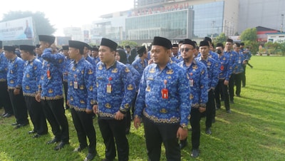 A group of people standing in lines on a grassy field wearing blue patterned uniforms and black hats. They appear to be organized in a formation and have ID badges on their chests. In the background, there is a large building with glass windows and a sign displaying 'RITA Super Mall'. Trees and other vegetation are visible along the perimeter.