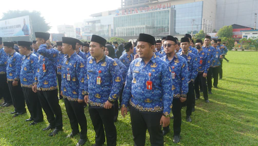 A group of people standing in lines on a grassy field wearing blue patterned uniforms and black hats. They appear to be organized in a formation and have ID badges on their chests. In the background, there is a large building with glass windows and a sign displaying 'RITA Super Mall'. Trees and other vegetation are visible along the perimeter.