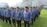 A group of people standing in lines on a grassy field wearing blue patterned uniforms and black hats. They appear to be organized in a formation and have ID badges on their chests. In the background, there is a large building with glass windows and a sign displaying 'RITA Super Mall'. Trees and other vegetation are visible along the perimeter.
