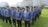 A group of people standing in lines on a grassy field wearing blue patterned uniforms and black hats. They appear to be organized in a formation and have ID badges on their chests. In the background, there is a large building with glass windows and a sign displaying 'RITA Super Mall'. Trees and other vegetation are visible along the perimeter.