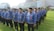 A group of people standing in lines on a grassy field wearing blue patterned uniforms and black hats. They appear to be organized in a formation and have ID badges on their chests. In the background, there is a large building with glass windows and a sign displaying 'RITA Super Mall'. Trees and other vegetation are visible along the perimeter.