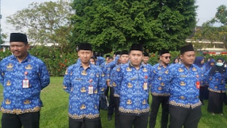 A group of people wearing matching blue shirts with gold embroidery patterns stand outdoors on a grassy area. They are mostly men, with a few women visible in the background. They appear to be in a formal or ceremonial setting, and many are wearing black hats and identification badges.