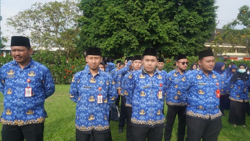 A group of people wearing matching blue shirts with gold embroidery patterns stand outdoors on a grassy area. They are mostly men, with a few women visible in the background. They appear to be in a formal or ceremonial setting, and many are wearing black hats and identification badges.