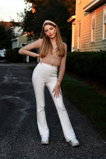 A young woman poses confidently on a residential street during what appears to be early evening with warm lighting. She wears a beige, long-sleeved top and white, high-waisted pants. Her hair is styled straight with a headband, and she's accessorized with necklaces and bracelets. Surrounding her are trees and a house side, suggesting a suburban environment.
