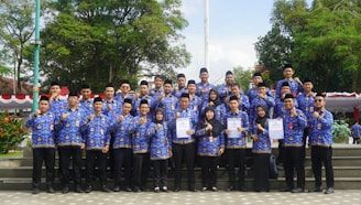 A large group of people, consisting mostly of men and a few women, stand on stone steps in an outdoor setting. They are wearing matching uniforms in blue with intricate patterns, and some are holding certificates. The background features lush green trees and a flagpole, adding to the formal and celebratory atmosphere.
