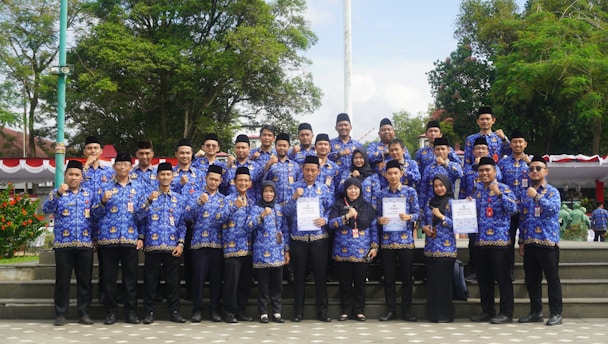 A large group of people, consisting mostly of men and a few women, stand on stone steps in an outdoor setting. They are wearing matching uniforms in blue with intricate patterns, and some are holding certificates. The background features lush green trees and a flagpole, adding to the formal and celebratory atmosphere.