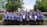 A large group of people, consisting mostly of men and a few women, stand on stone steps in an outdoor setting. They are wearing matching uniforms in blue with intricate patterns, and some are holding certificates. The background features lush green trees and a flagpole, adding to the formal and celebratory atmosphere.