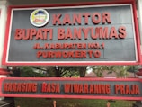 A large sign with bold red letters displaying the name 'KANTOR BUPATI BANYUMAS' and an address 'JL. KABUPATEN NO.1 PURWOKERTO'. Above the text is an emblem with detailed design elements. The background is a dark gray color, and the sign is bordered with a red frame. It is positioned outdoors, with trees and foliage partially visible behind it.