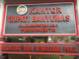 A large sign with bold red letters displaying the name 'KANTOR BUPATI BANYUMAS' and an address 'JL. KABUPATEN NO.1 PURWOKERTO'. Above the text is an emblem with detailed design elements. The background is a dark gray color, and the sign is bordered with a red frame. It is positioned outdoors, with trees and foliage partially visible behind it.