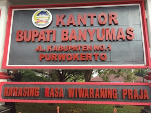 A large sign with bold red letters displaying the name 'KANTOR BUPATI BANYUMAS' and an address 'JL. KABUPATEN NO.1 PURWOKERTO'. Above the text is an emblem with detailed design elements. The background is a dark gray color, and the sign is bordered with a red frame. It is positioned outdoors, with trees and foliage partially visible behind it.