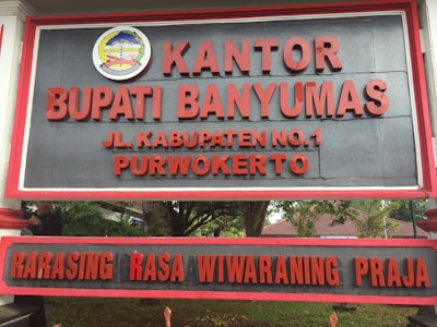A large sign with bold red letters displaying the name 'KANTOR BUPATI BANYUMAS' and an address 'JL. KABUPATEN NO.1 PURWOKERTO'. Above the text is an emblem with detailed design elements. The background is a dark gray color, and the sign is bordered with a red frame. It is positioned outdoors, with trees and foliage partially visible behind it.