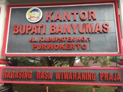A large sign with bold red letters displaying the name 'KANTOR BUPATI BANYUMAS' and an address 'JL. KABUPATEN NO.1 PURWOKERTO'. Above the text is an emblem with detailed design elements. The background is a dark gray color, and the sign is bordered with a red frame. It is positioned outdoors, with trees and foliage partially visible behind it.