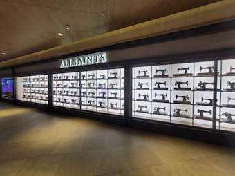 Displayed within a large glass case is a well-organized collection of vintage sewing machines, arranged in a grid fashion. Above the display case, a bright sign with illuminated letters reads 'ALLSAINTS'. The backdrop is brightly lit, contrasting with the dark silhouettes of the sewing machines. The area around the display features a modern design with a wooden ceiling and tiled floor.