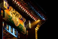 Ornate architectural details of a traditional structure featuring intricate paintings and colorful patterns under a dimly lit canopy. The artwork includes depictions of landscapes and floral motifs, highlighted by a warm light that contrasts with the dark, nighttime background.