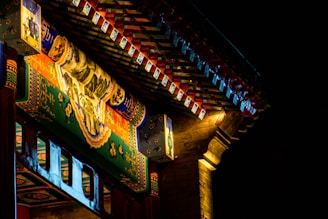 Ornate architectural details of a traditional structure featuring intricate paintings and colorful patterns under a dimly lit canopy. The artwork includes depictions of landscapes and floral motifs, highlighted by a warm light that contrasts with the dark, nighttime background.