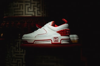 A pair of stylish sneakers with a sleek design featuring white leather and red accents is displayed against a dark, textured background. The shoes have a modern, sporty appearance with a unique pattern of red spikes on the heel.