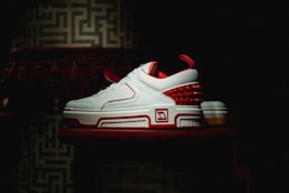 A pair of stylish sneakers with a sleek design featuring white leather and red accents is displayed against a dark, textured background. The shoes have a modern, sporty appearance with a unique pattern of red spikes on the heel.