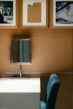 A modern vanity setup featuring a rectangular mirror on a wooden wall above a white desk. Three framed artworks hang on the wall, consisting of a black-and-white fashion photograph and two images of gemstones or jewelry designs. A blue chair is placed in front of the desk.