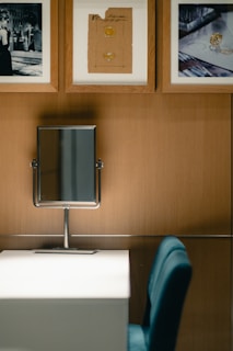 A modern vanity setup featuring a rectangular mirror on a wooden wall above a white desk. Three framed artworks hang on the wall, consisting of a black-and-white fashion photograph and two images of gemstones or jewelry designs. A blue chair is placed in front of the desk.